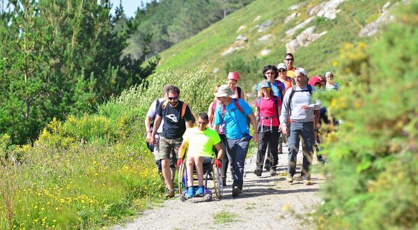 Grupo de pessoas a caminhar numa trilha, ajudando um jovem numa cadeira de rodas.