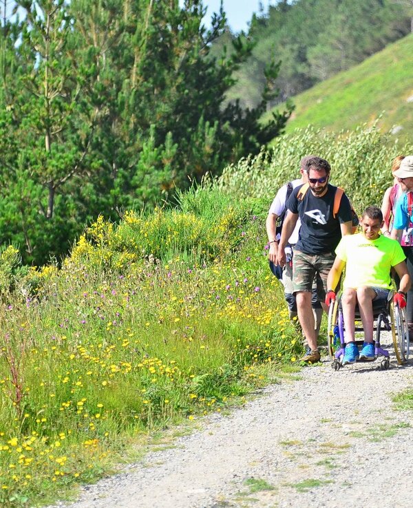 Grupo de pessoas a caminhar numa trilha, ajudando um jovem numa cadeira de rodas.