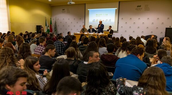 Grande sala cheia de estudantes a assistir a uma apresentação em painel.