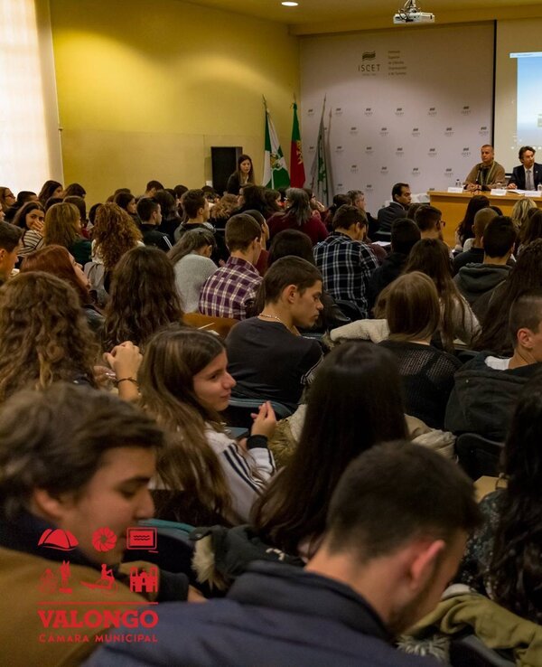Grande sala cheia de estudantes a assistir a uma apresentação em painel.
