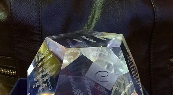 Um troféu em cristal com gravações em várias faces, dentro de uma caixa azul.