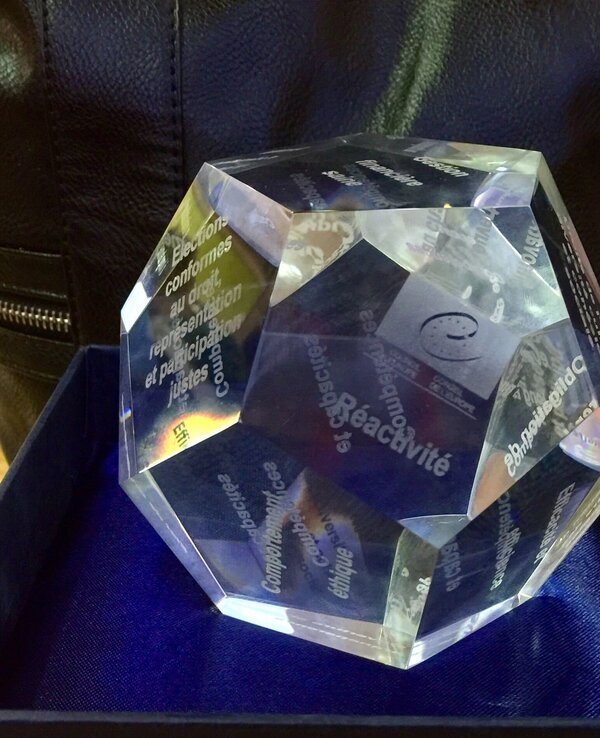 Um troféu em cristal com gravações em várias faces, dentro de uma caixa azul.