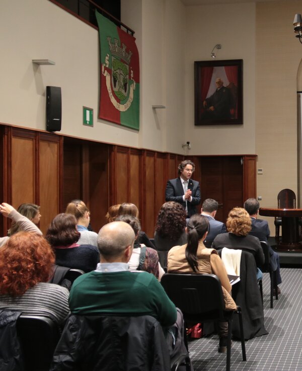 Reunião com orador em palco, público atento e apresentação ao fundo.