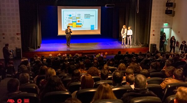 Público assistindo a uma apresentação num auditório, com um orador e dois assistentes.