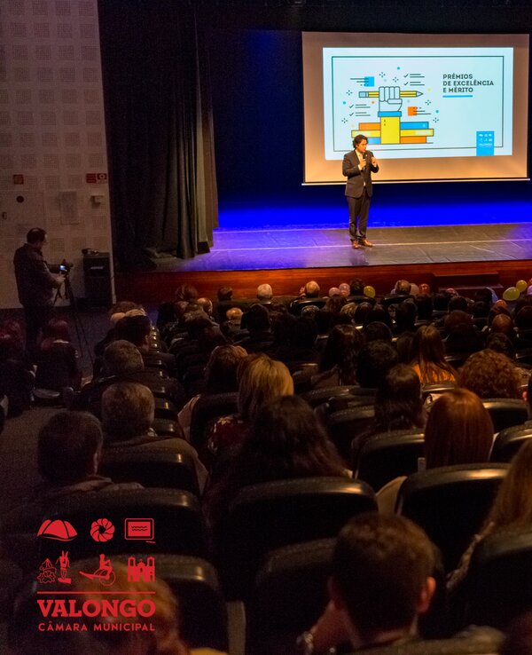 Público assistindo a uma apresentação num auditório, com um orador e dois assistentes.