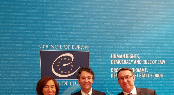 Três pessoas seguram um certificado em frente a um fundo azul do Conselho da Europa.