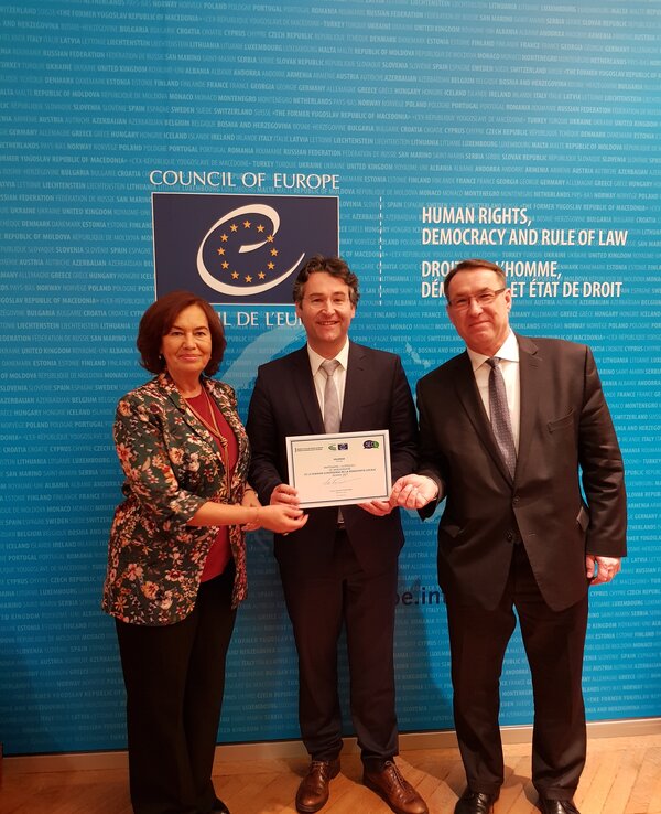 Três pessoas seguram um certificado em frente a um fundo azul do Conselho da Europa.