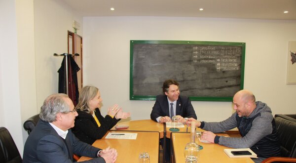 Reunião em mesa de madeira com quatro pessoas a discutir, quadro negro ao fundo.