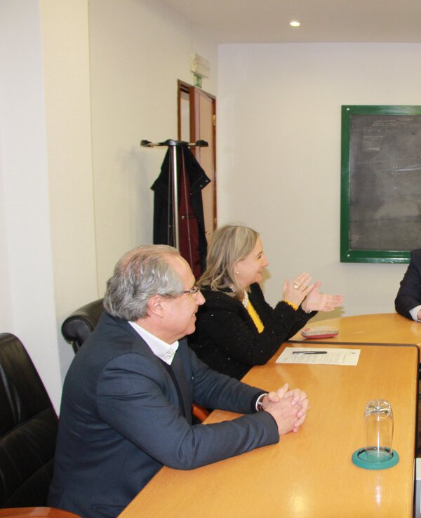 Reunião em mesa de madeira com quatro pessoas a discutir, quadro negro ao fundo.