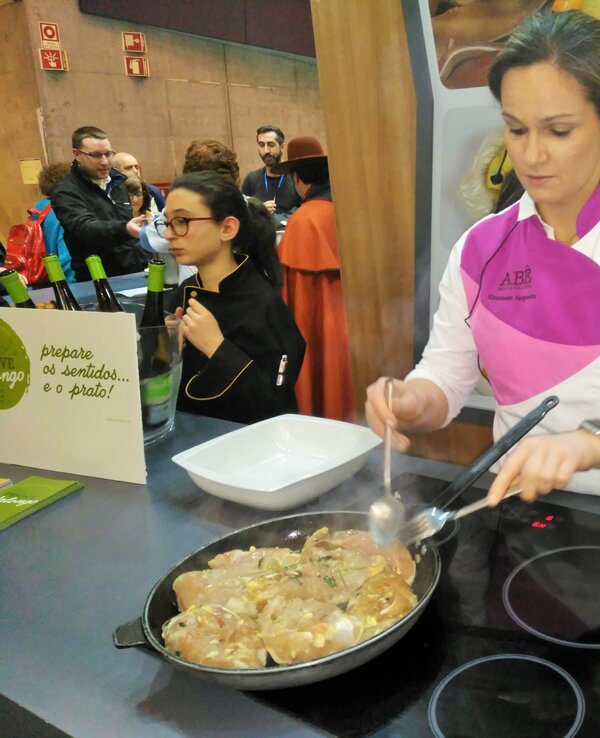 Uma chef a cozinhar carne numa frigideira, com pessoas a observar ao fundo.