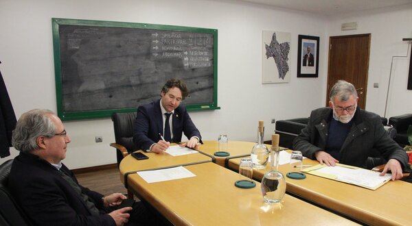 Reunião em sala com três homens à mesa, um a escrever e quadro negro ao fundo.