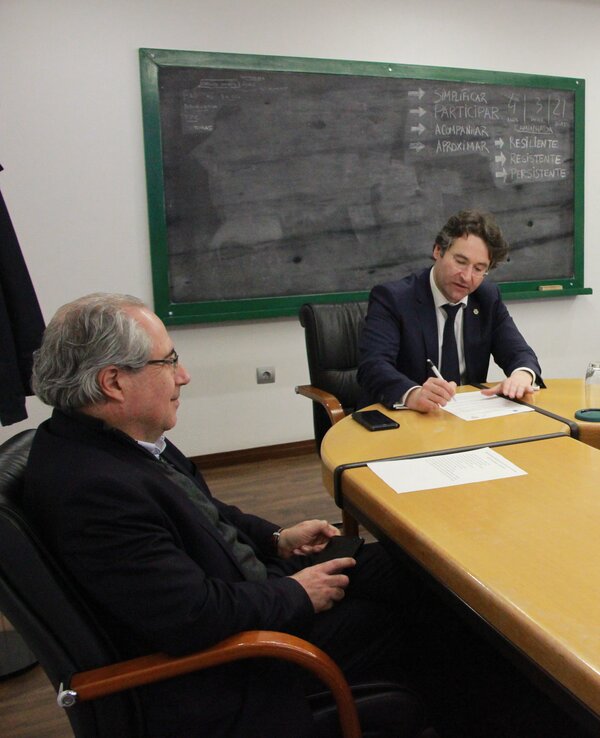 Reunião em sala com três homens à mesa, um a escrever e quadro negro ao fundo.