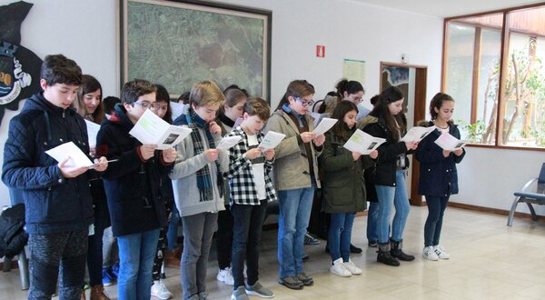 Grupo de jovens a cantar, segurando folhas com letras, em ambiente interior.