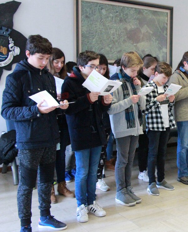 Grupo de jovens a cantar, segurando folhas com letras, em ambiente interior.