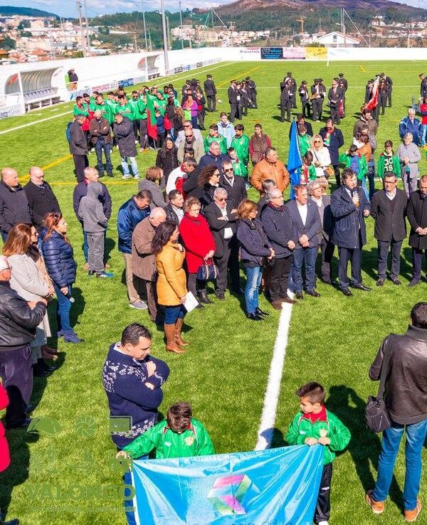 Grupo de pessoas reunidas num campo de futebol, com bandeiras e uniformes.