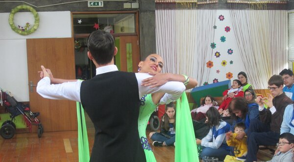 Casal a dançar numa sala, com crianças a assistir e decoração colorida ao fundo.