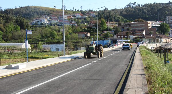 Estrada rural com um trator e um carro, rodeada por casas e vegetação.