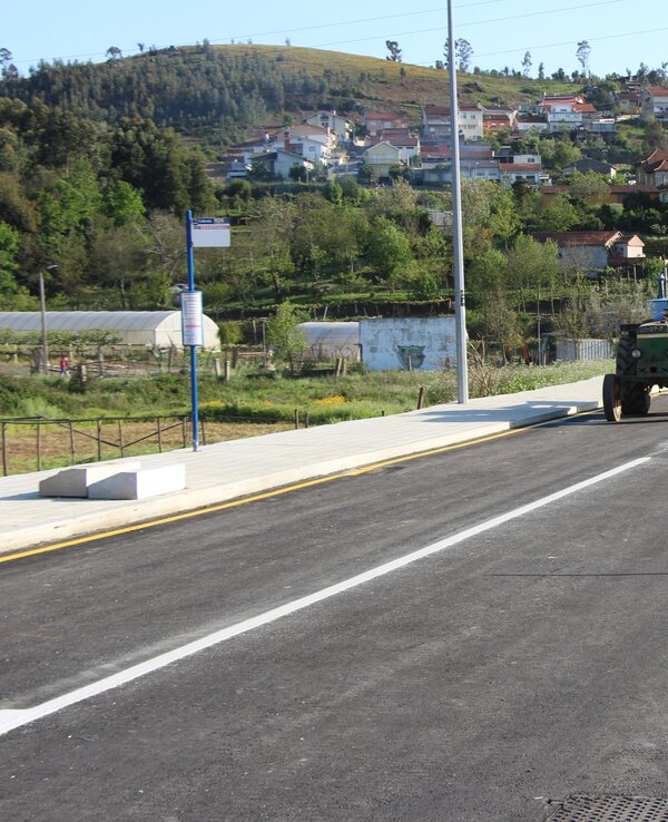Estrada rural com um trator e um carro, rodeada por casas e vegetação.