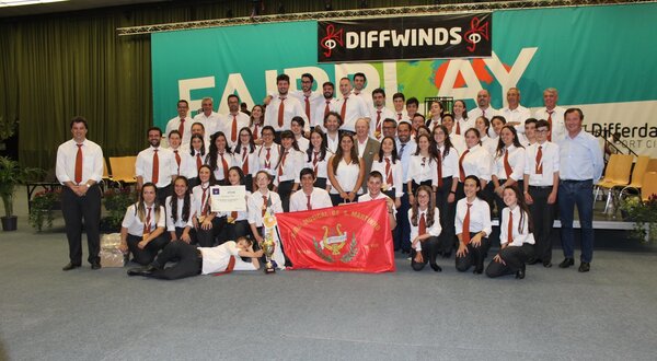 Grupo grande de músicos em palco, com bandeira e certificado de participação.
