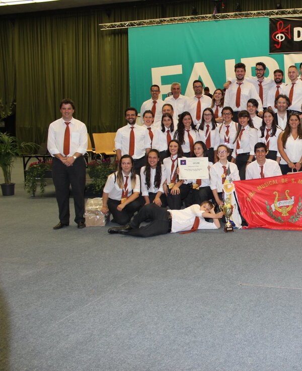 Grupo grande de músicos em palco, com bandeira e certificado de participação.