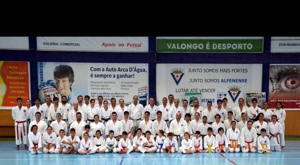 Grupo de praticantes de karaté em uniforme branco, posando em um pavilhão desportivo.