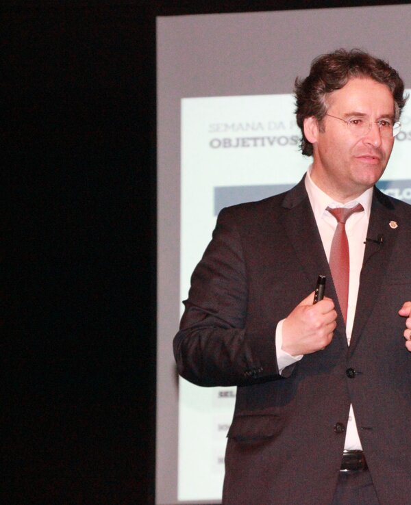 Homem em fato a apresentar num evento, com diapositivo ao fundo.