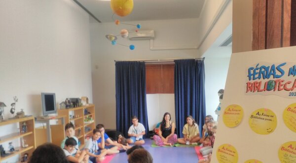 Grupo de crianças sentadas em círculo numa sala de atividades, com cartazes.