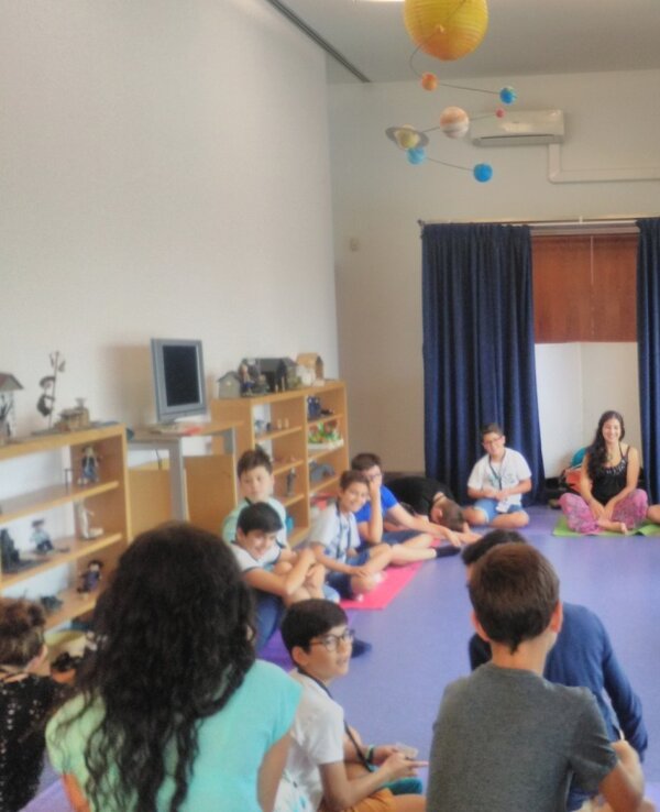 Grupo de crianças sentadas em círculo numa sala de atividades, com cartazes.